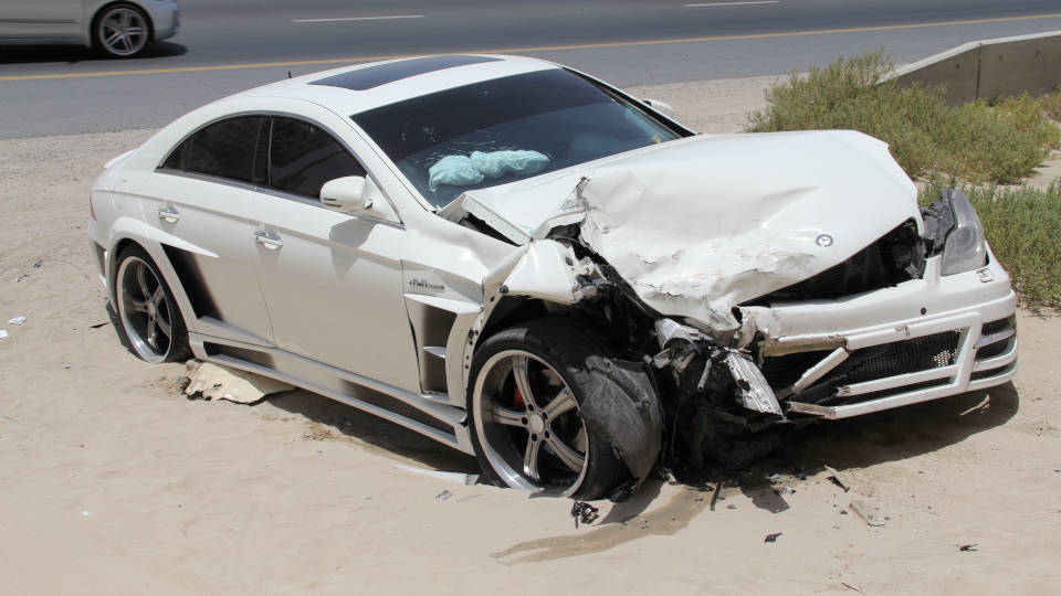 A picture of a brand new Mercedes Benz sitting in sand off to the side of the highway with serious front damage.