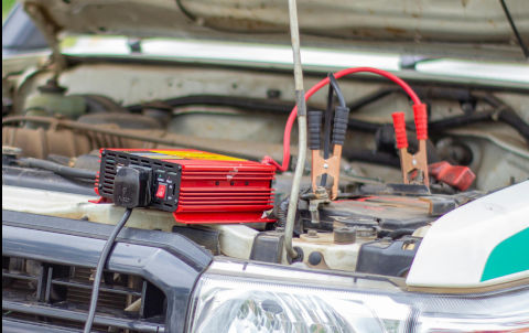 A picture of a car battery charger sitting on top of the engine, hood open, plugged into the battery charging it.